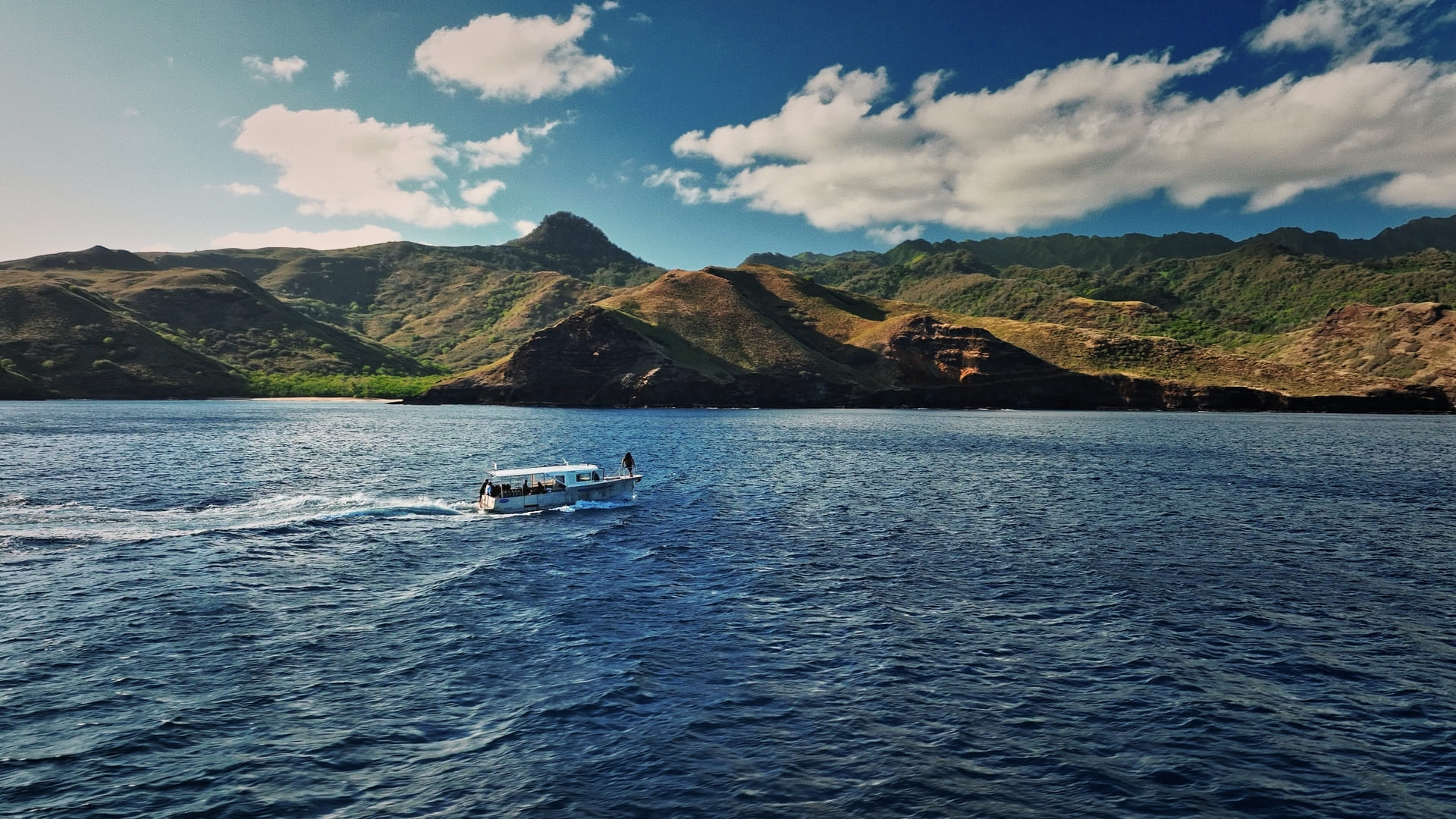 Petit bateau naviguant sur une mer bleue devant des montagnes verdoyantes sous un ciel partiellement nuageux.
